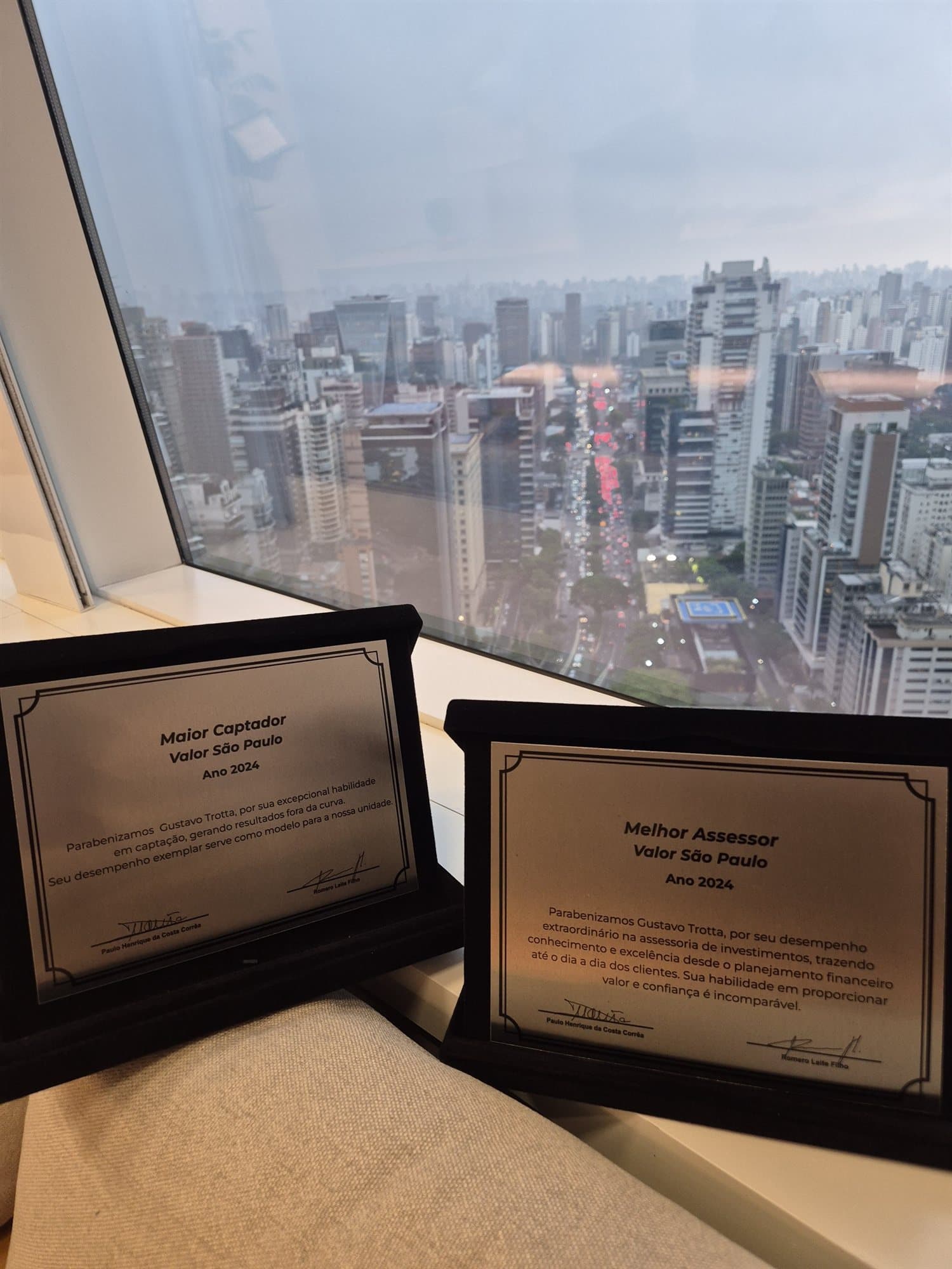 Placas dos prêmios Melhor Assessor e Maior Captador da unidade São Paulo da Valor Investimentos, ano 2024, com a vista da cidade de São Paulo ao fundo